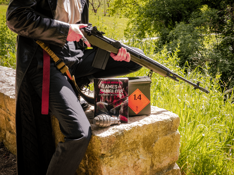 Photo promotionnelle de lames et sanglots : la chanson de l'assassin, le livre figure entre un assassin au manteau noir tenant un fusil et une caisse de munitions
